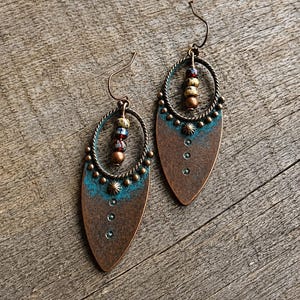 May include: A pair of teardrop-shaped copper earrings featuring a turquoise patina finish at the top, a twisted wire oval frame, and a vertical row of three small embedded crystals. Each earring includes a central dangling strand of multi-colored beads.