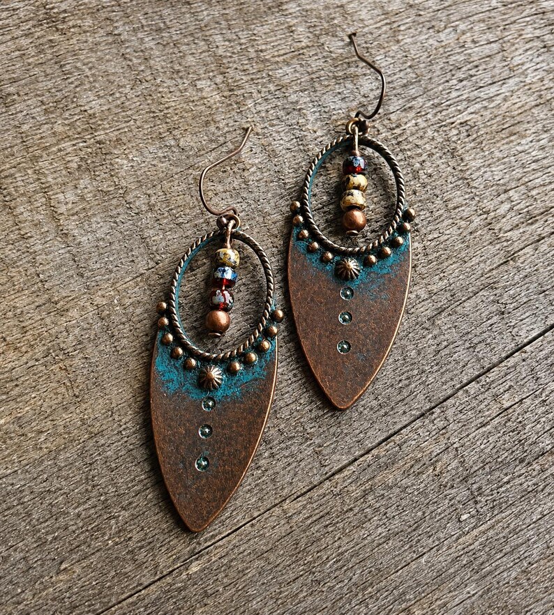 May include: A pair of teardrop-shaped copper earrings featuring a turquoise patina finish at the top, a twisted wire oval frame, and a vertical row of three small embedded crystals. Each earring includes a central dangling strand of multi-colored beads.