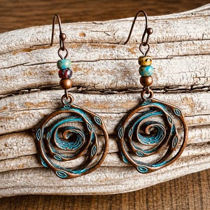 May include: Copper-coloured earrings with a spiral design. The earrings have a turquoise and copper colour scheme, with a spiral centre and small beaded accents. The earrings are displayed on a piece of driftwood.