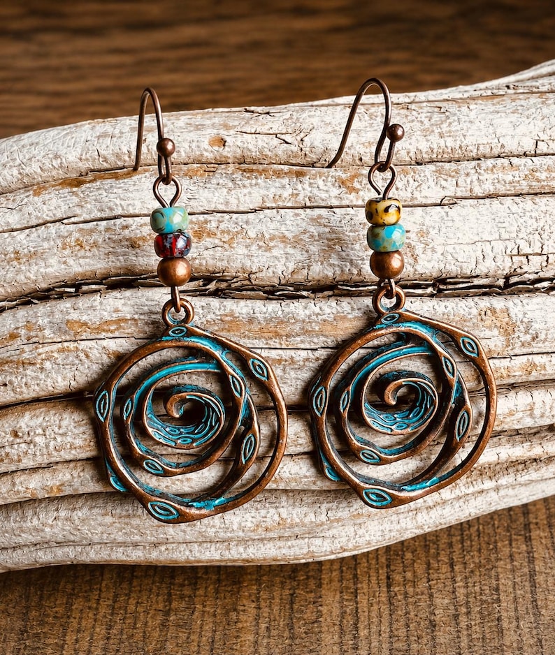 May include: Copper-coloured earrings with a spiral design. The earrings have a turquoise and copper colour scheme, with a spiral centre and small beaded accents. The earrings are displayed on a piece of driftwood.