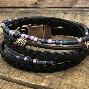 May include: A black leather bracelet with a braided design and multiple strands of iridescent purple and blue beads. The bracelet has two silver metal accents and a magnetic clasp.