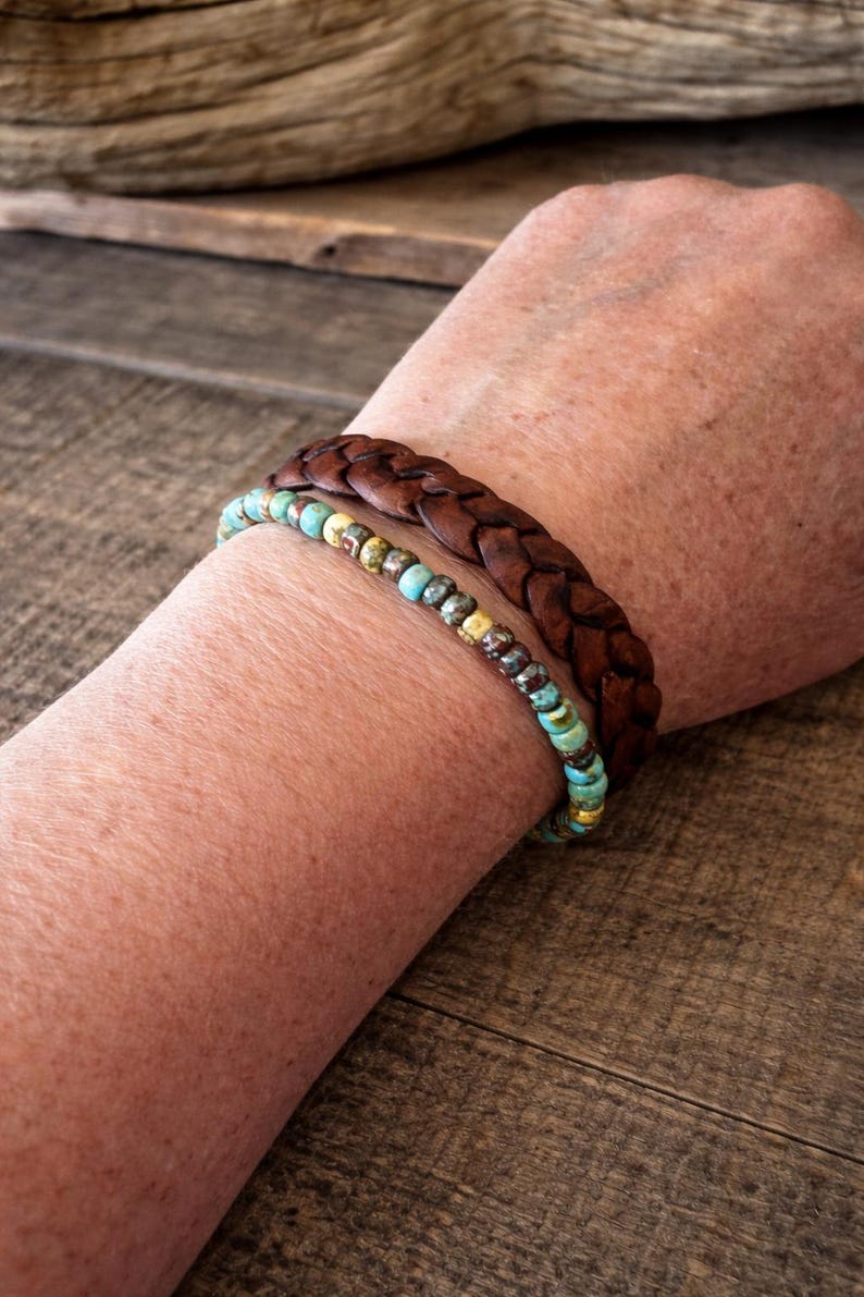 Two bracelets are shown. The top bracelet is a dark brown, braided leather band. The bottom bracelet is made of small, round beads in shades of turquoise, yellow, and brown. The bracelets are worn on a person's arm.