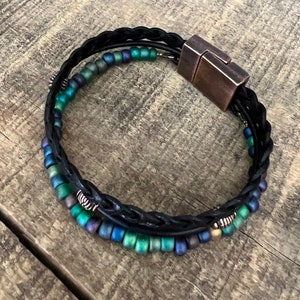 Puede incluir: Un brazalete de cuero negro con un dise&ntilde;o trenzado y un cierre magn&eacute;tico. El brazalete est&aacute; adornado con cuentas iridiscentes verdes y azules.