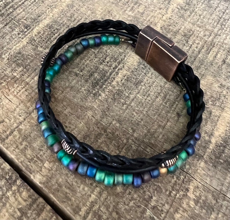 Puede incluir: Un brazalete de cuero negro con un dise&ntilde;o trenzado y un cierre magn&eacute;tico. El brazalete est&aacute; adornado con cuentas iridiscentes verdes y azules.