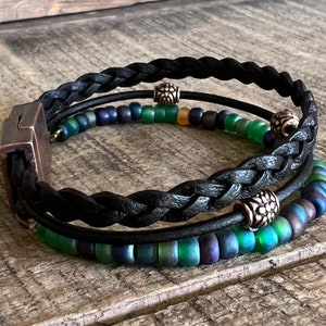Puede incluir: Un brazalete trenzado de cuero negro con tres hileras de cuentas iridiscentes verdes y azules y detalles plateados.