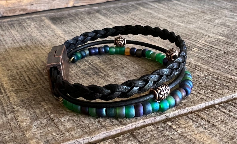 Puede incluir: Un brazalete trenzado de cuero negro con tres hileras de cuentas iridiscentes verdes y azules y detalles plateados.