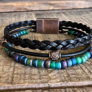 Puede incluir: Un brazalete de cuero negro con un dise&ntilde;o trenzado y un cierre magn&eacute;tico de cobre. El brazalete tiene dos filas de cuentas iridiscentes verdes y azules.