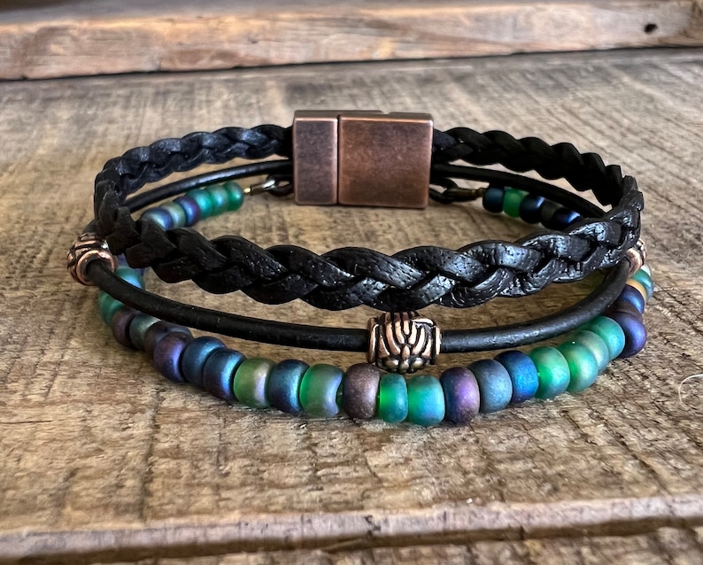 Puede incluir: Un brazalete de cuero negro con un dise&ntilde;o trenzado y un cierre magn&eacute;tico de cobre. El brazalete tiene dos filas de cuentas iridiscentes verdes y azules.
