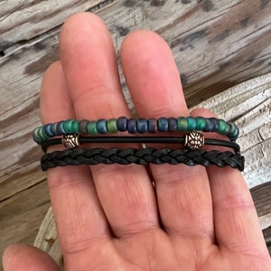 Puede incluir: Un brazalete de capas con una banda de cuero trenzado negro y una hebra de cuentas iridiscentes verdes y azules. El brazalete tiene dos cuentas de metal plateado con un dise&ntilde;o decorativo.