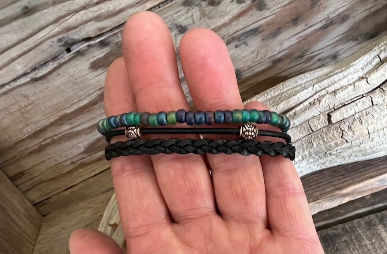 Puede incluir: Un brazalete de capas con una banda de cuero trenzado negro y una hebra de cuentas iridiscentes verdes y azules. El brazalete tiene dos cuentas de metal plateado con un dise&ntilde;o decorativo.