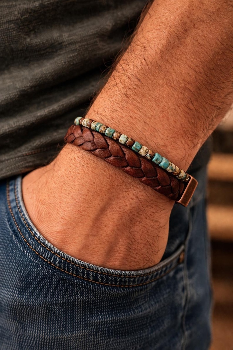 Two bracelets on a wrist. The bottom bracelet is braided brown leather with a copper clasp. The top bracelet has small turquoise and brown beads. The person is wearing a gray shirt and blue jeans.
