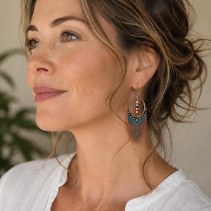 May include: A dangle earring featuring a circular copper frame with a central vertical row of three small multicolored beads. Below the circle is a pointed, shield-shaped copper pendant with a teal blue patina finish and a small star detail.
