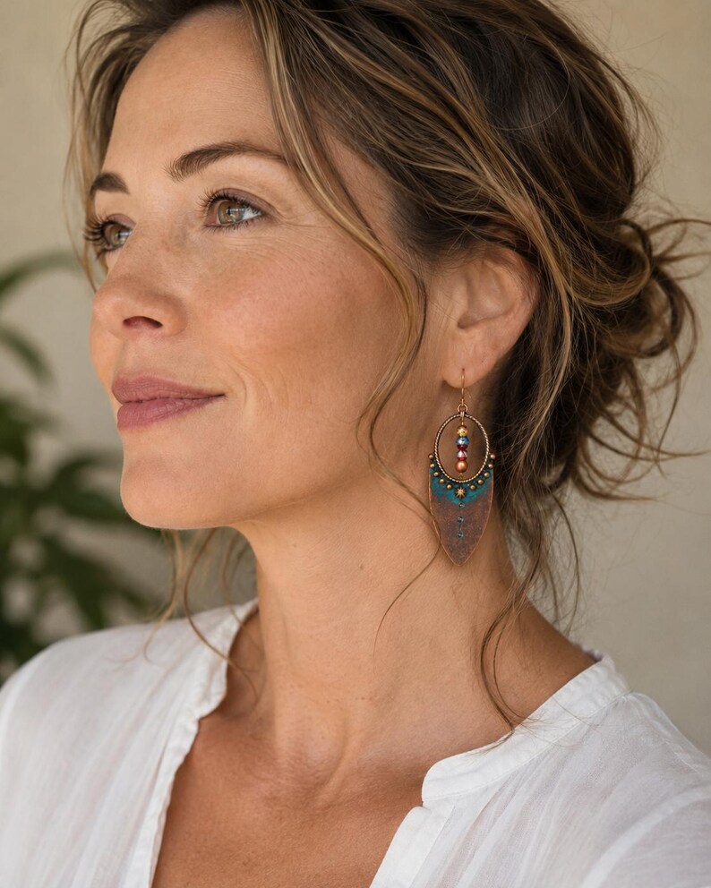 May include: A dangle earring featuring a circular copper frame with a central vertical row of three small multicolored beads. Below the circle is a pointed, shield-shaped copper pendant with a teal blue patina finish and a small star detail.