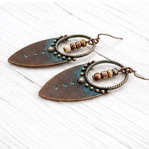 May include: A pair of dangle earrings featuring a pointed, teardrop-shaped copper-toned metal base with a dark patina and teal accents. Each earring includes a circular twisted wire frame holding three small, multi-colored patterned beads.