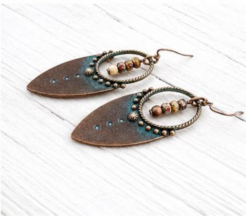 May include: A pair of dangle earrings featuring a pointed, teardrop-shaped copper-toned metal base with a dark patina and teal accents. Each earring includes a circular twisted wire frame holding three small, multi-colored patterned beads.