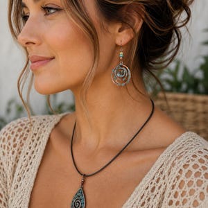 May include: A woman wearing a necklace and earrings. The necklace features a teardrop pendant with a turquoise design. The earrings are circular with a spiral design and turquoise accents. The jewellery is set against a cream-coloured top.
