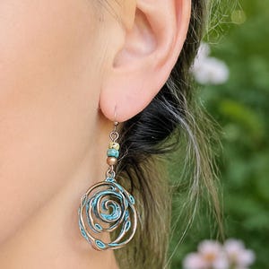 May include: A close-up of a copper-coloured earring with a spiral design and turquoise accents. The earring is suspended from a small hook and features small, colourful beads. The background is blurred, with green and white tones.