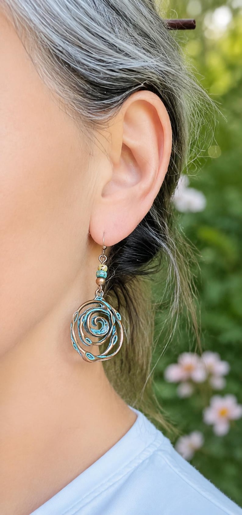 May include: A close-up of a copper-coloured earring with a spiral design and turquoise accents. The earring is suspended from a small hook and features small, colourful beads. The background is blurred, with green and white tones.