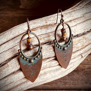 May include: A pair of copper-toned dangle earrings featuring a twisted circular frame attached to a pointed shield-shaped base. The metal has a weathered patina with teal accents, small decorative studs, and a vertical row of three tiny dark beads.