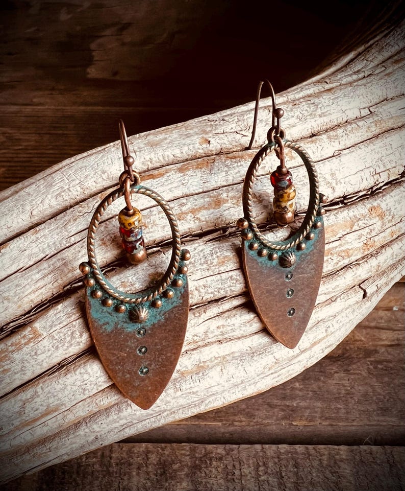 May include: A pair of copper-toned dangle earrings featuring a twisted circular frame attached to a pointed shield-shaped base. The metal has a weathered patina with teal accents, small decorative studs, and a vertical row of three tiny dark beads.