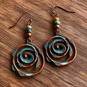 May include: A pair of copper-toned earrings featuring a spiral design with a turquoise blue centre. The earrings are adorned with small beads in green, brown, and yellow. They are presented on a wooden surface.