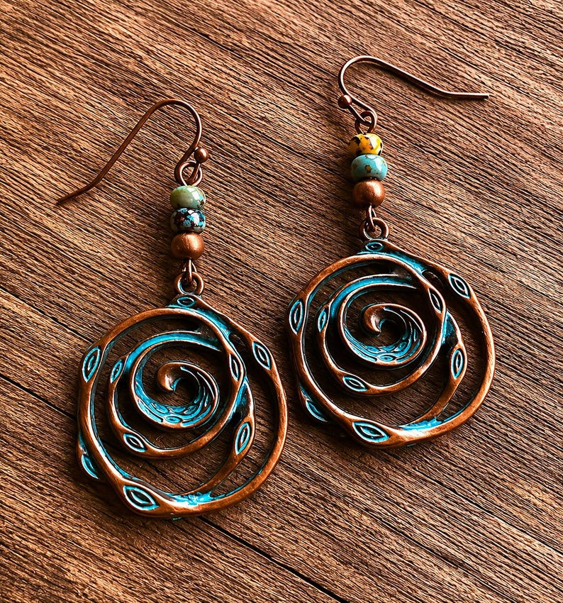 May include: A pair of copper-toned earrings featuring a spiral design with a turquoise blue centre. The earrings are adorned with small beads in green, brown, and yellow. They are presented on a wooden surface.