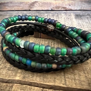 May include: A black leather braided bracelet with three strands of iridescent green glass beads. The bracelet has a silver bead accent.