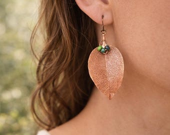 May include: Copper-colored leaf earrings with a textured surface and small green and blue beads. The earrings are hanging from a copper-colored hook. The earrings are a nature-inspired accessory.