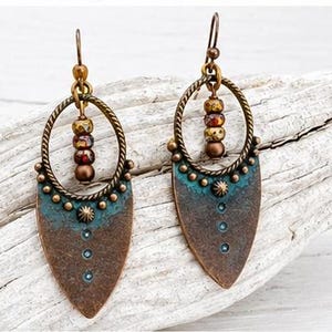 May include: A pair of dangle earrings featuring a bronze-toned metal shield shape with a teal patina finish. Each earring has a twisted circular frame holding three small, multi-colored beads and a central metal stud with a dot pattern.
