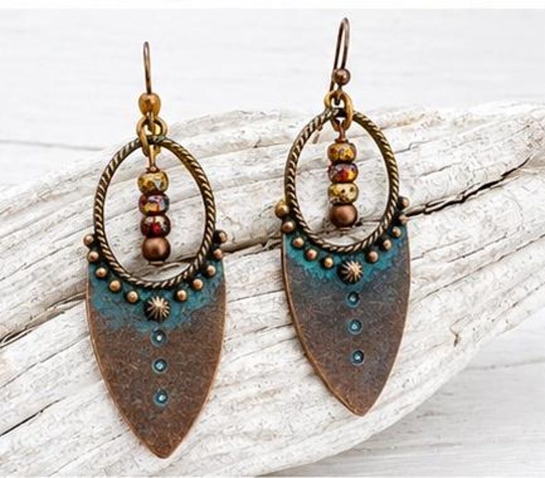May include: A pair of dangle earrings featuring a bronze-toned metal shield shape with a teal patina finish. Each earring has a twisted circular frame holding three small, multi-colored beads and a central metal stud with a dot pattern.