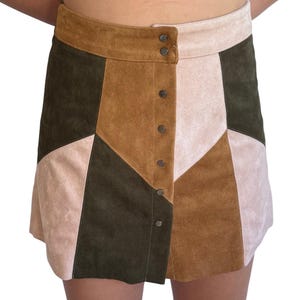 Może przedstawiać: Mini spódnica zamszowa w stylu patchwork w odcieniach brązu, oliwkowej zieleni i różu. Spódnica ma zapinany na guziki przód i wysoką talię. Spódnica jest w stylu retro.
