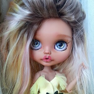 Può includere: Una bambola con lunghi capelli biondi e grandi occhi azzurri. Indossa un vestito giallo con un sopraabito di pizzo.