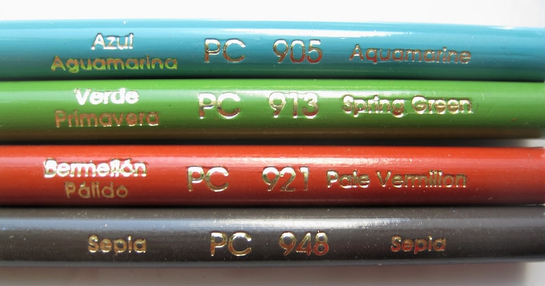 Op de afbeelding: Vier kleurpotloden met gouden letters. De potloden zijn gelabeld met "Azul Aguamarina PC 905 Aquamarine", "Verde Primavera PC 913 Spring Green", "Bermell&oacute;n P&aacute;lido PC 921 Pale Vermilion" en "Sepia PC 948 Sepia".