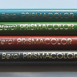Op de afbeelding: Vier Prismacolor kleurpotloden met etiketten in het Spaans en Engels. De potloden zijn gelabeld "Azul  Azuliano PC 905", "Verde Primavera PC 913", "Bermellon Brillo PC 921", en "Sepia PC 942".