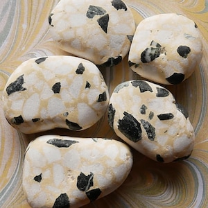 May include: Five smooth, rounded stones with a speckled pattern of white, beige, and black. The stones are arranged on a swirled, yellow and beige background. The stones vary in size and shape.