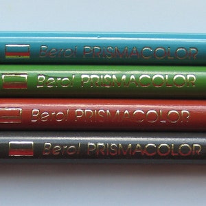 Op de afbeelding: Vijf Prismacolor kleurpotloden met de merknaam en "Made in Mexico" erop gedrukt. De potloden zijn blauw, groen, rood, grijs en zwart. De kleurennamen en -nummers zijn op elk potlood gedrukt: "Azul Aguamarina PC 105", "Verde Primavera PC 010", "Bermellon PC 921", "Gris PC 948" en "Negro PC 949".