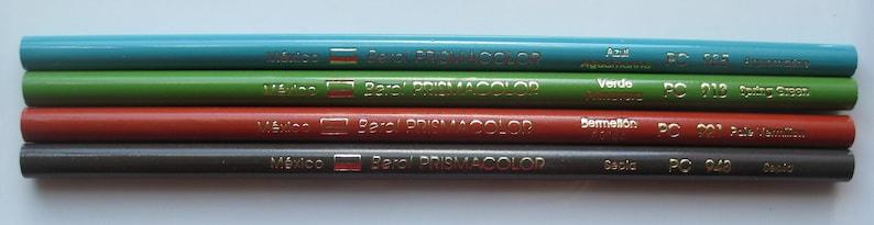 Op de afbeelding: Vijf Prismacolor kleurpotloden met de merknaam en "Made in Mexico" erop gedrukt. De potloden zijn blauw, groen, rood, grijs en zwart. De kleurennamen en -nummers zijn op elk potlood gedrukt: "Azul Aguamarina PC 105", "Verde Primavera PC 010", "Bermellon PC 921", "Gris PC 948" en "Negro PC 949".