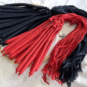 May include: Close-up of bundles of red and black rope. The red rope is tightly wound and has a tassel at the end. The black rope is braided. The ropes are arranged on a wooden embroidery hoop.