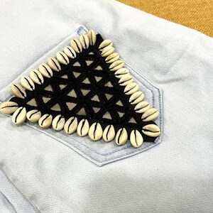 May include: Light blue denim pocket with a triangular black crochet design, edged with white seashells. The pocket is part of a light blue denim garment, likely jeans or shorts. The design adds a unique, beachy aesthetic.