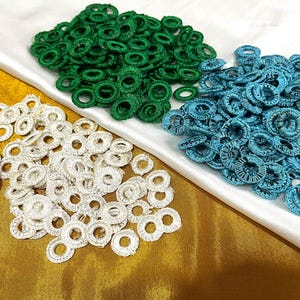 May include: An assortment of handmade lace rings in three colors: white, green, and teal. The rings are arranged in separate piles on a white and gold fabric background. The lace rings are likely for crafting or embellishment.