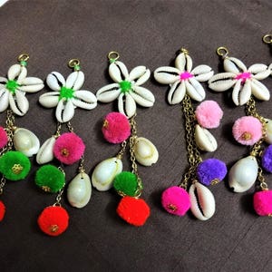 Peut inclure: Cinq breloques en forme de fleur en coquille blanche avec des chaînes dorées et des pompons colorés. Les breloques sont suspendues à un fond en tissu brun.