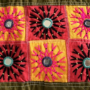 Może przedstawiać: Kolorowa ścienna wisząca z tkaniny w stylu patchwork. Każdy kwadrat ma inny haft z okrągłym lustrem w środku. Kolory to czerwony, żółty, różowy, czarny i zielony.