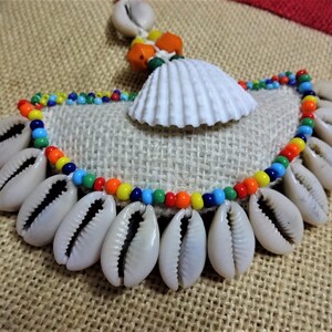 May include: A colorful beaded necklace with cowrie shells and a white seashell pendant. The necklace is on a burlap background.