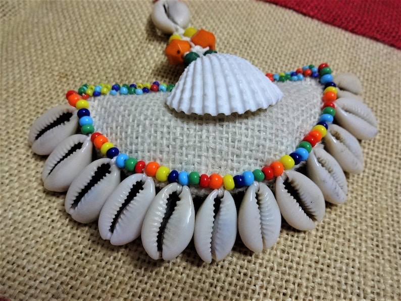 May include: A colorful beaded necklace with cowrie shells and a white seashell pendant. The necklace is on a burlap background.