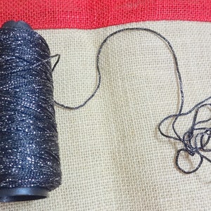 Puede incluir: Una bobina de hilo negro con purpurina plateada, con un tramo de hilo desenrollado y que se encuentra sobre una superficie de arpillera.