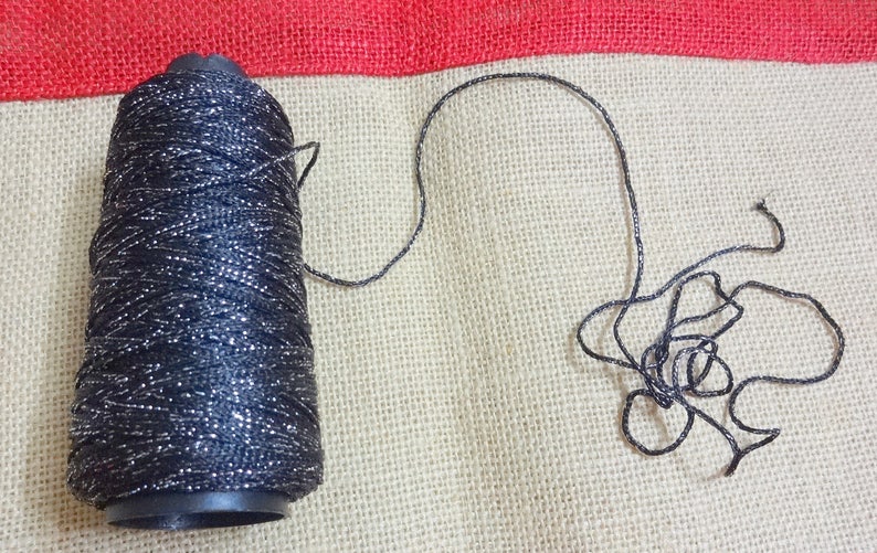 Puede incluir: Una bobina de hilo negro con purpurina plateada, con un tramo de hilo desenrollado y que se encuentra sobre una superficie de arpillera.