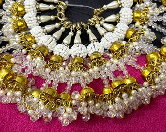 May include: A close-up of a decorative trim with gold and white beads. The trim features small, round, gold-colored beads with a dangling crystal bead chain.