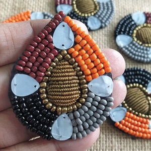 May include: A set of four beaded teardrop-shaped embellishments with a central gold circle surrounded by black, orange, gray, and maroon beads. The embellishments have a white, pearlescent finish.