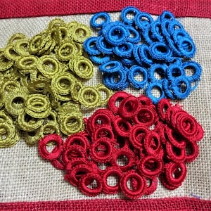 Puede incluir: Una colección de pequeños anillos de ganchillo en tres colores: dorado, azul y rojo. Los anillos están dispuestos en forma de corazón sobre un fondo de arpillera.