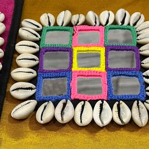 May include: Close-up of a decorative square featuring small mirrors framed with colorful crochet, surrounded by white seashells. The crochet frames are in shades of green, purple, yellow, pink, and blue. The background is a golden-yellow color.